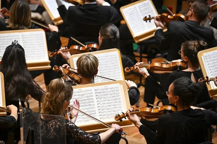 Overhead view of a symphony orchestra during rehearsal or performance, with musicians in black holding violins and sheet music on music stands.