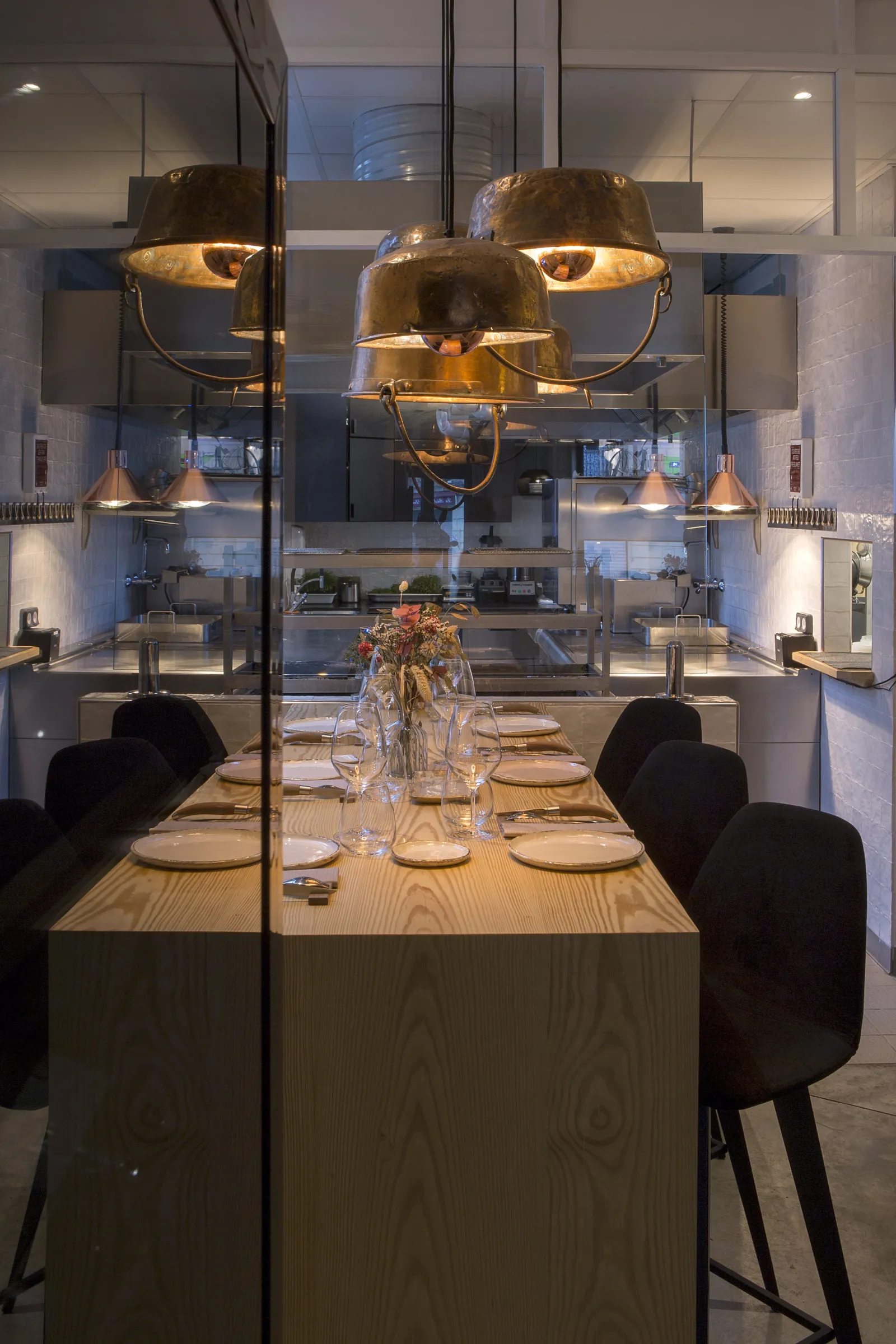 Modern dining room with light wooden table, black chairs, suspended golden pendant lights, glass partitions and open kitchen visible in the background, located at Cent33 in Bordeaux.