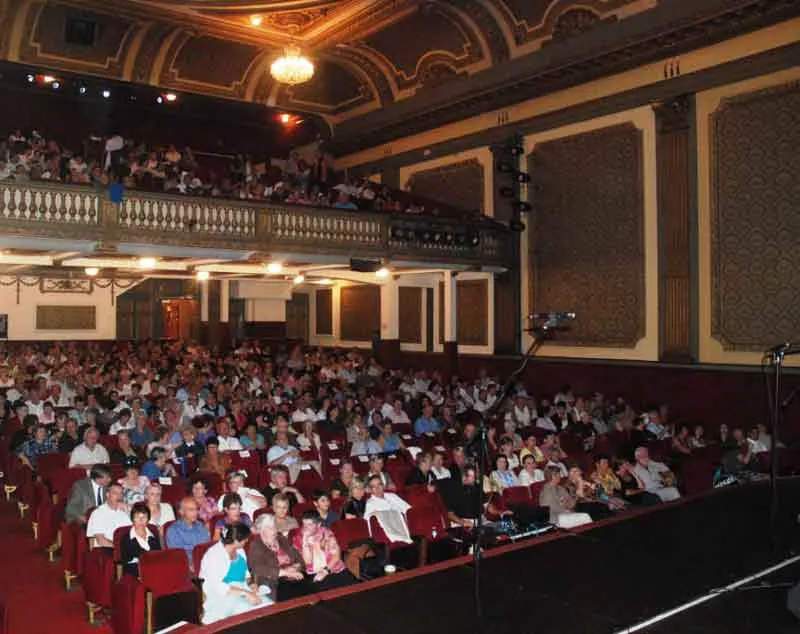 Interior of a Bordeaux theater with audience seated in red chairs, decorated balconies, ornate gilt ceiling and paneled walls.