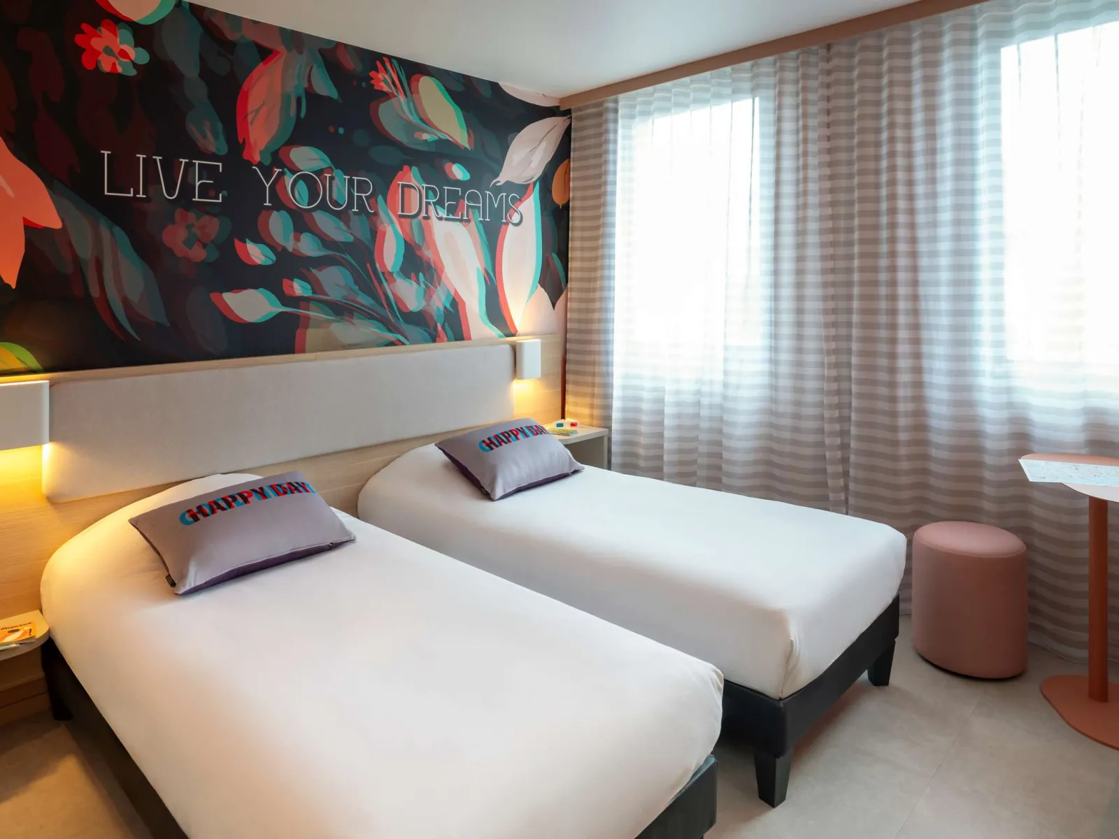 Modern hotel room featuring two single beds, vibrant mural wall art with 'Live Your Dreams' text, gray and white checkered curtains, contemporary furnishings and warm lighting.