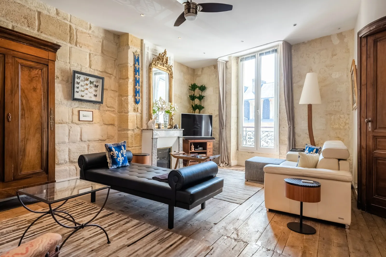Elegant living room in a Haussmann apartment in Bordeaux featuring exposed stone walls, gilt fireplace, contemporary black and beige furniture, large windows overlooking the street, and refined decor.
