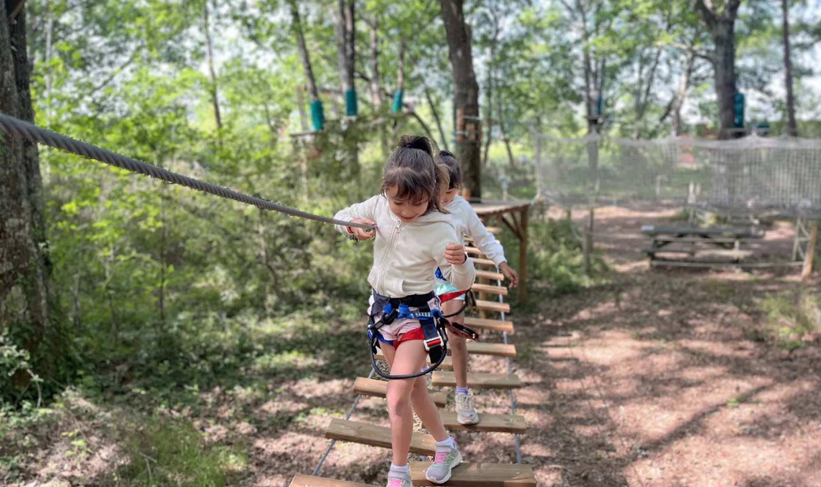 A child wearing a safety harness crosses an aerial obstacle course in the forest, walking on suspended wooden boards between trees at Tépacap Bordeaux-Mérignac park.