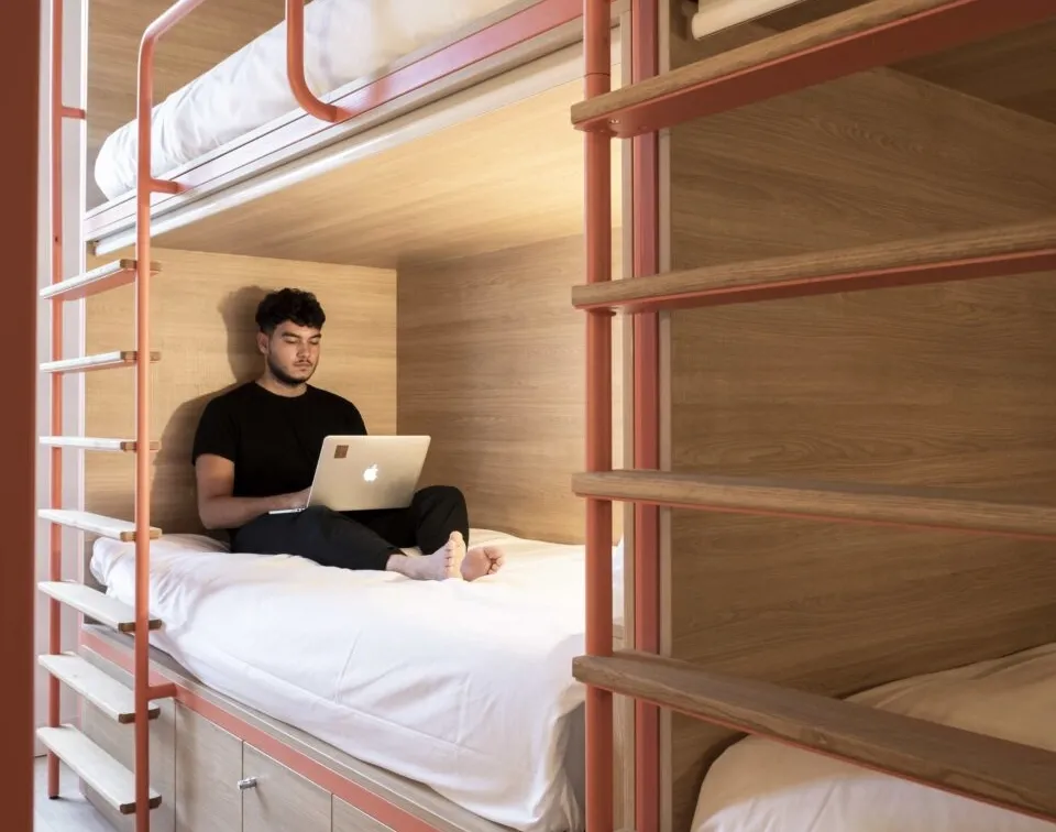 A young man sitting on a bunk bed in a hostel room in Bordeaux, working on a MacBook laptop.