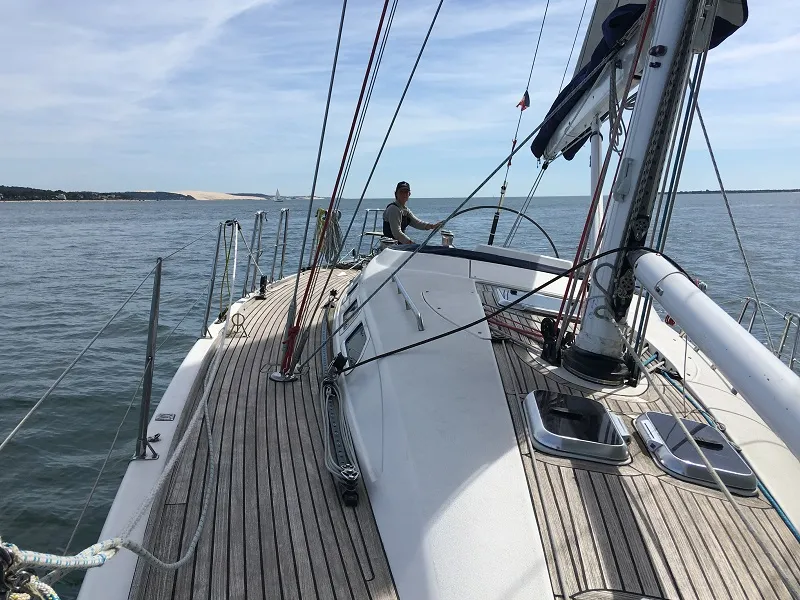 Deck view of a white sailing yacht with a sailor at the helm sailing on calm waters, coastal landscape in the background near Bordeaux