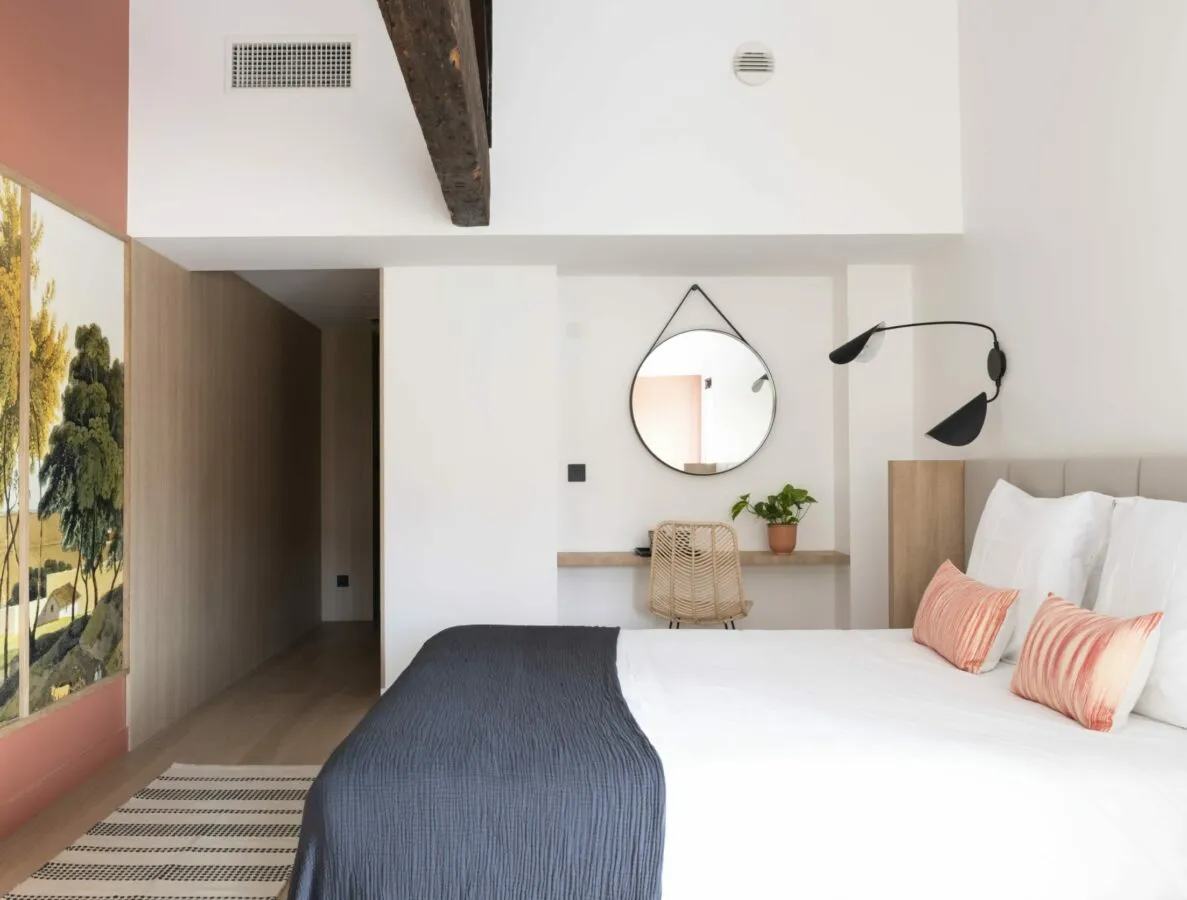 Modern hostel room in Bordeaux featuring a white bed with coral pillows, round mirror, wooden desk, potted plant, and botanical wall art