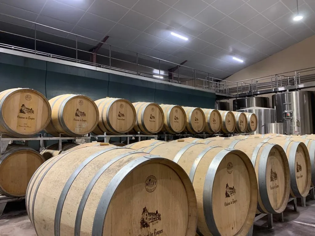 Interior of Château de Roques winery in Bordeaux featuring rows of wooden oak barrels arranged for wine aging, with stainless steel equipment visible in the background