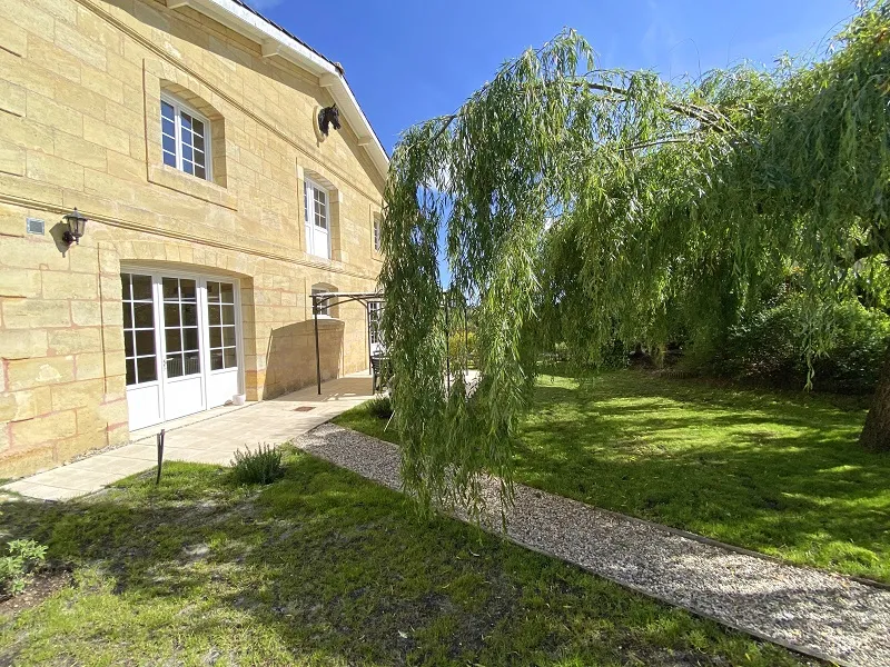 Stone house facade with paved terrace, white French doors and weeping willow in a sunny green garden