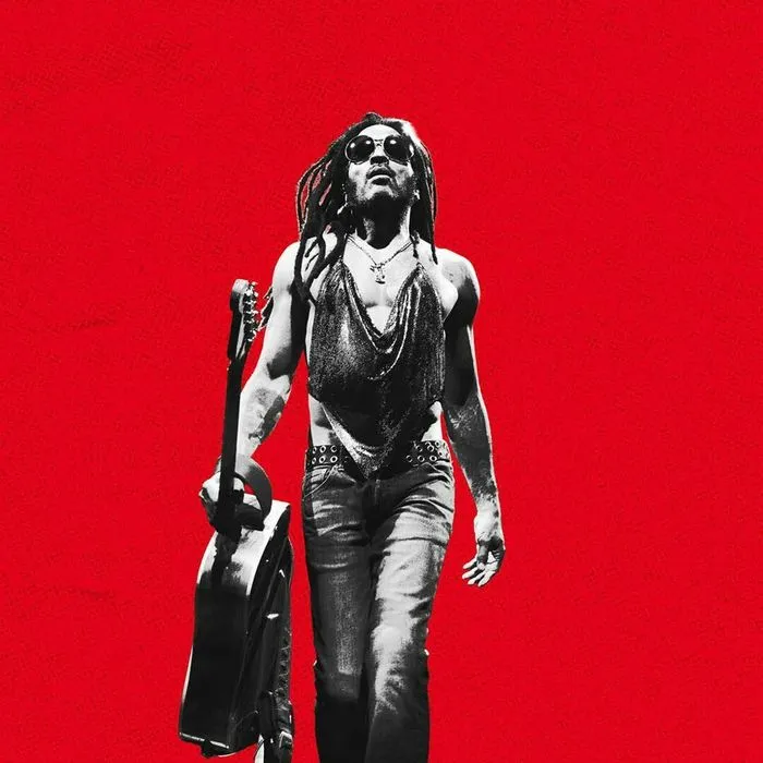 Lenny Kravitz performing at Arkéa Arena in Bordeaux, wearing sunglasses and holding a guitar against a bright red background.