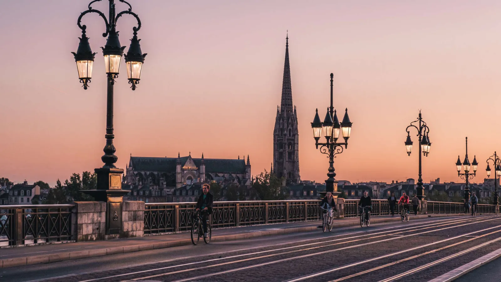 Stone Bridge in Bordeaux at sunset with Saint-Michel Spire in the background, cyclists crossing the illuminated bridge with ornamental street lamps.