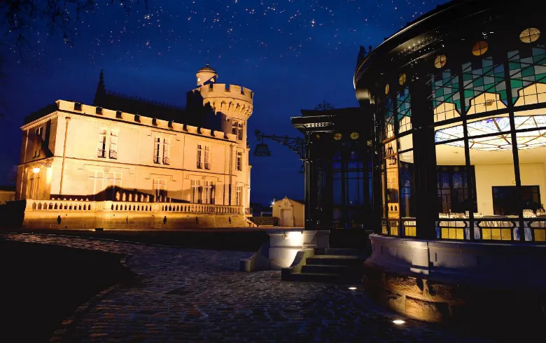 Château Pape Clément lit up at night in Bordeaux, featuring its distinctive square tower and stone facades reflected in surrounding water beneath a starry sky.