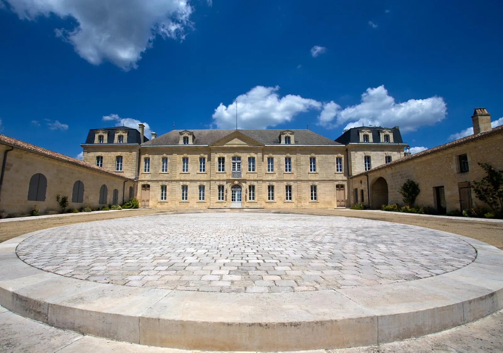 Inner courtyard of Château Soutard in Bordeaux featuring a stone mansion with mansard roof, circular stone paving, and colonnaded wings under clear blue sky.