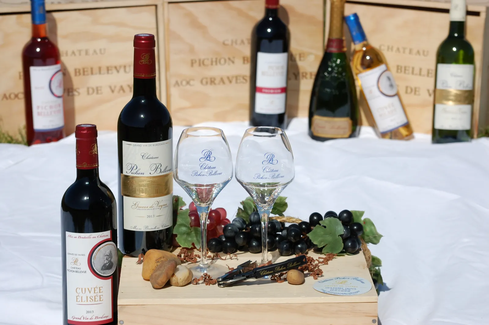 Wine tasting display from Château Pichon Bellevue in Bordeaux featuring wine glasses with red and white wines, black grapes, bread, and wine bottles in the background on wooden crates.