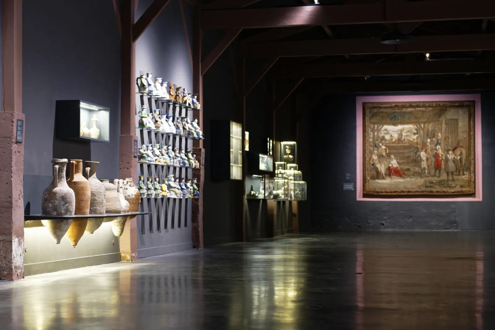 Museum gallery displaying ancient artifacts: ceramics and amphorae in an illuminated display case on the left, collection of figurines in a central glass case, and large framed historical fresco on the right wall, within an industrial architecture with ex
