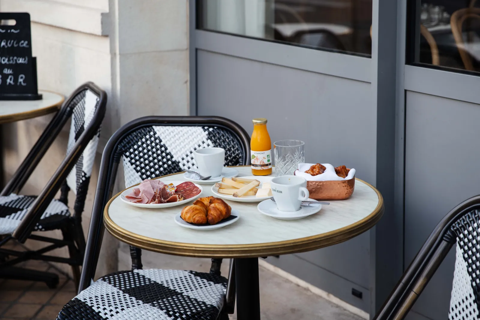 Round bistro table set with French breakfast featuring croissants, cured meats, cheese, fries, coffee and orange juice, black and white wicker chairs, outdoor seating at Bistrot Le Saint-Jean in Bordeaux.