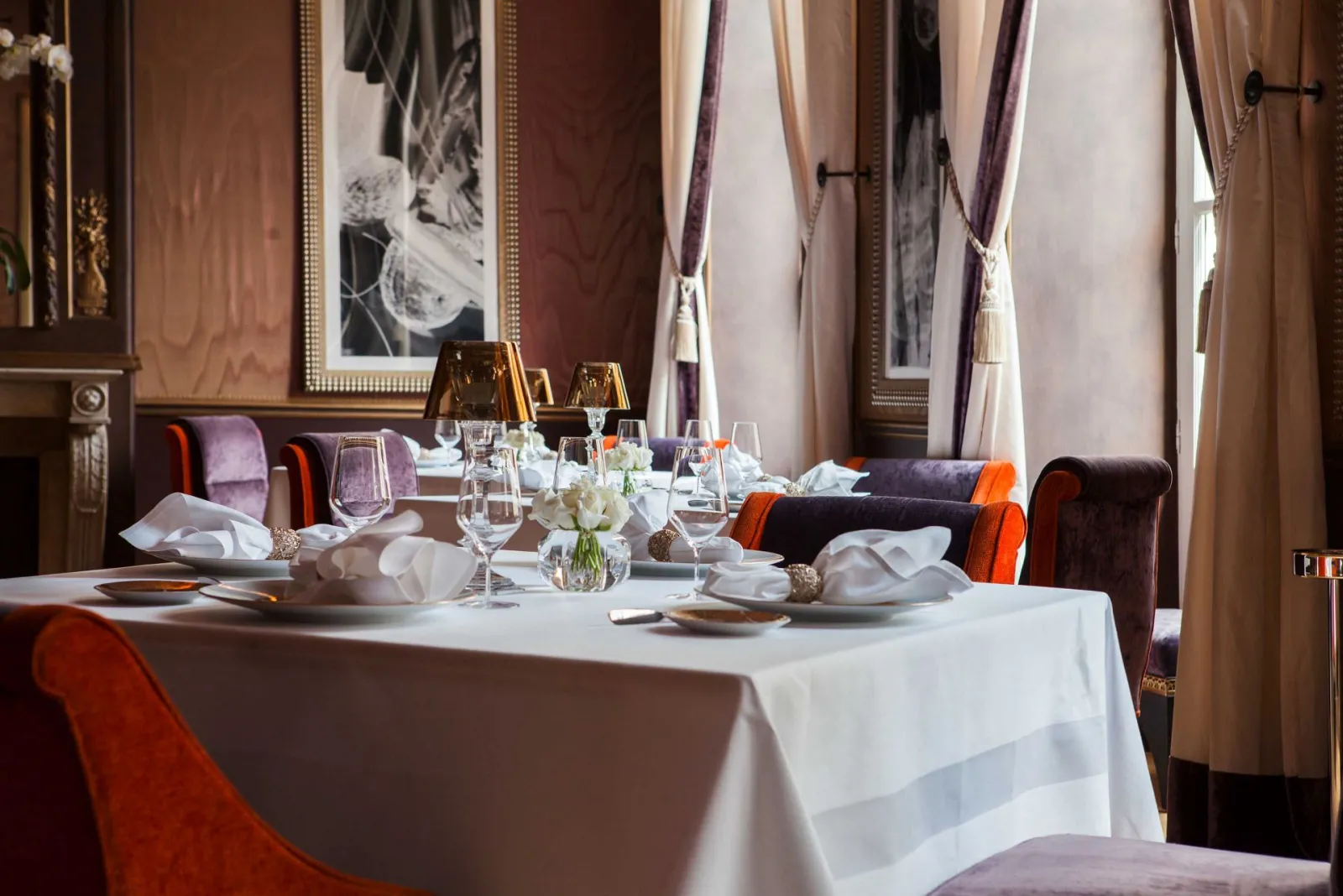 Elegant fine dining restaurant room with round table setting, burgundy and orange upholstered chairs, fine glassware, white linens and sophisticated drapes at Le Pressoir d'Argent Gordon Ramsay in Bordeaux.