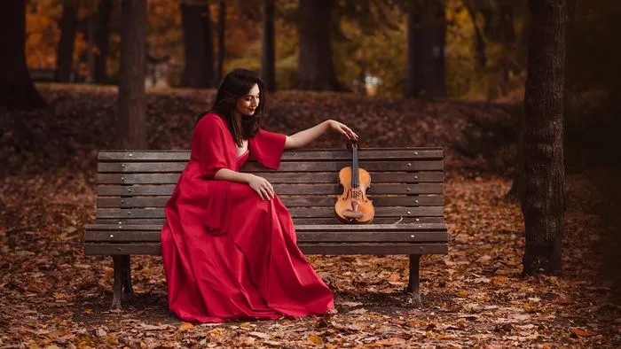 Woman in a red dress sitting on a bench in autumn, holding a violin in a golden leaf forest in Bordeaux