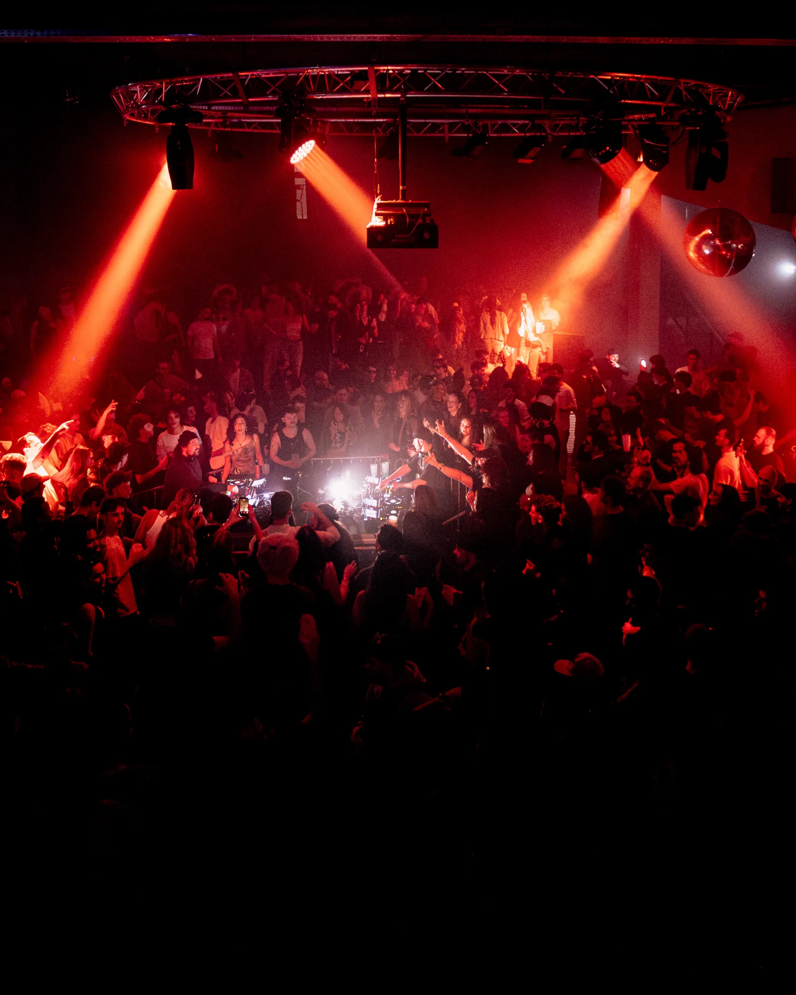 Crowd of concert-goers in a Bordeaux venue, lit by intense red stage lights from the metal ceiling structure, creating a festive and energetic atmosphere.