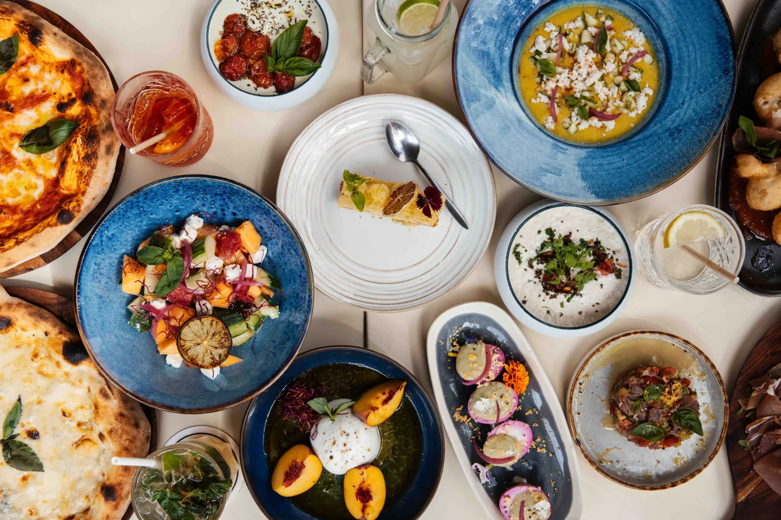 Restaurant table featuring gourmet dishes including pizzas, curries, tartares and colorful accompaniments, with rosé wine glasses and water, in a modern fine dining setting.
