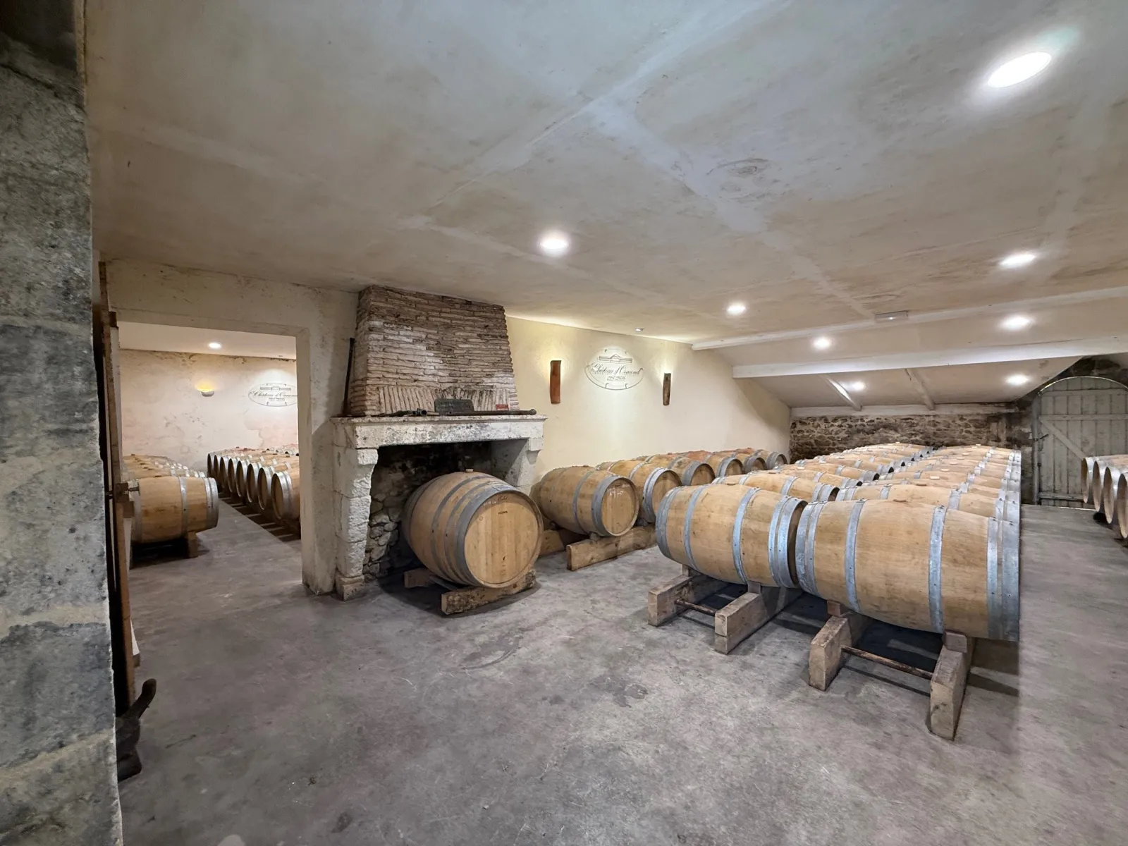 Interior of a wine cellar featuring wooden oak barrels on wooden stands, a stone fireplace, recessed lighting, and doorways leading to additional storage rooms at Château d'Osmond in Bordeaux.