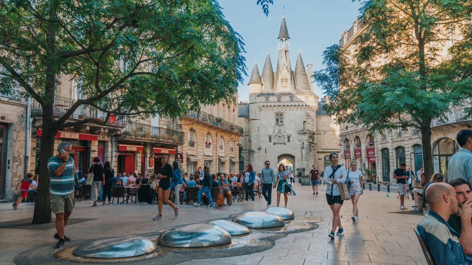 Cailhau Gate, a historic monument in Bordeaux, seen from a shaded public square with visitors, cafés, and ground sculptures