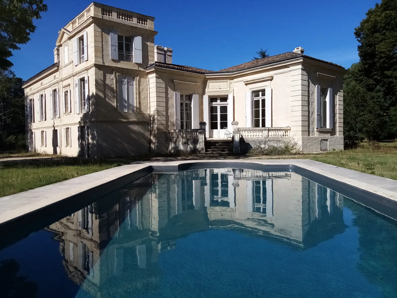 Mayne La Merci Castle in Bordeaux featuring a white stone neoclassical mansion with a square tower, reflected in a modern swimming pool in the foreground.