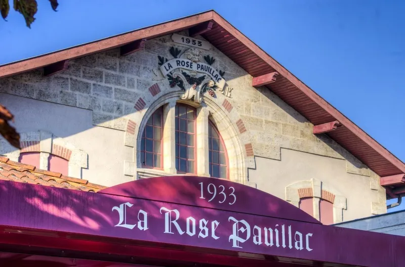 Front facade of La Rose Pauillac wine cellar in Bordeaux, built in 1933, featuring stone architecture, arched windows, and a purple storefront sign.