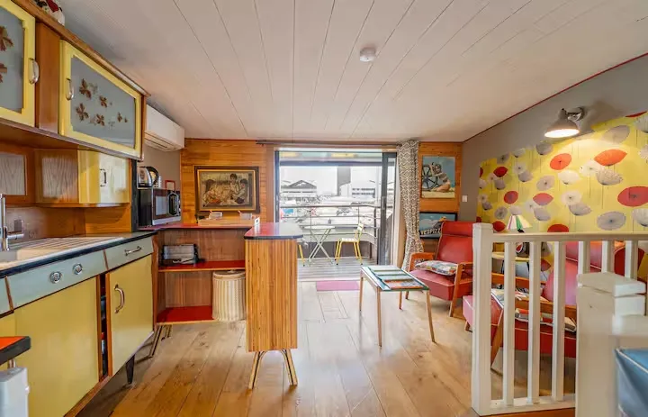 Colorful interior of a converted barge featuring a vintage yellow kitchen, retro-style living area, children's space with white crib and decorated walls, overlooking the Garonne River in Bordeaux