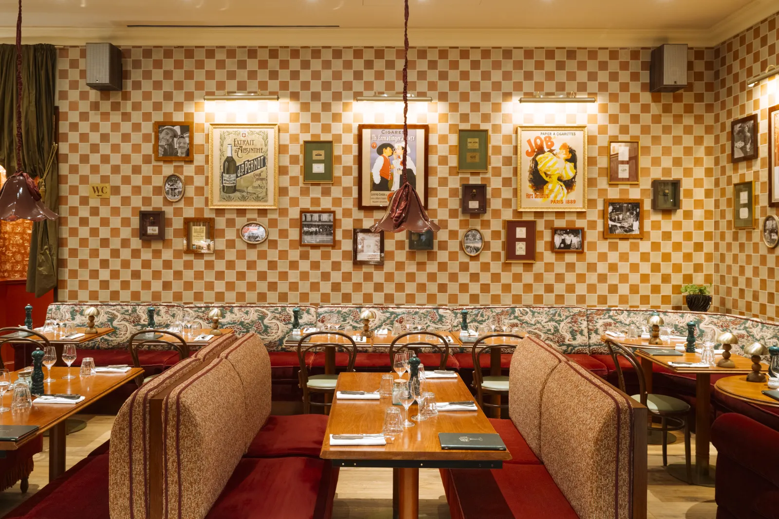 Elegant interior of Brasserie Vinatier in Bordeaux featuring red banquettes, wooden tables, checkered beige and brown walls, and a collection of vintage posters and historical framed artwork.