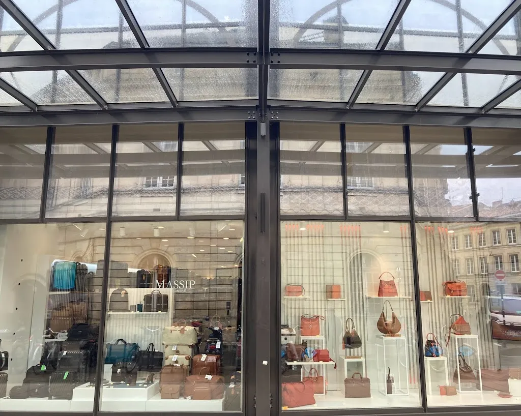 Storefront of Massip Maroquinerie in Bordeaux displaying leather handbags and accessories in various colors through glass windows beneath an architectural glass canopy.
