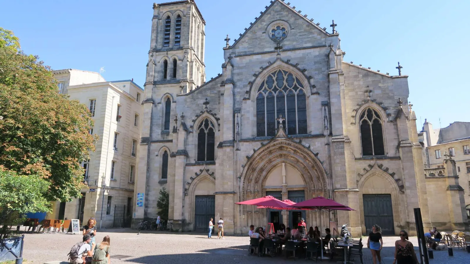 Front view of Saint-Pierre Church in Bordeaux, a Gothic stone building featuring a square bell tower, ornate pointed arch windows, and rose windows, with people relaxing in the plaza below under red umbrellas.
