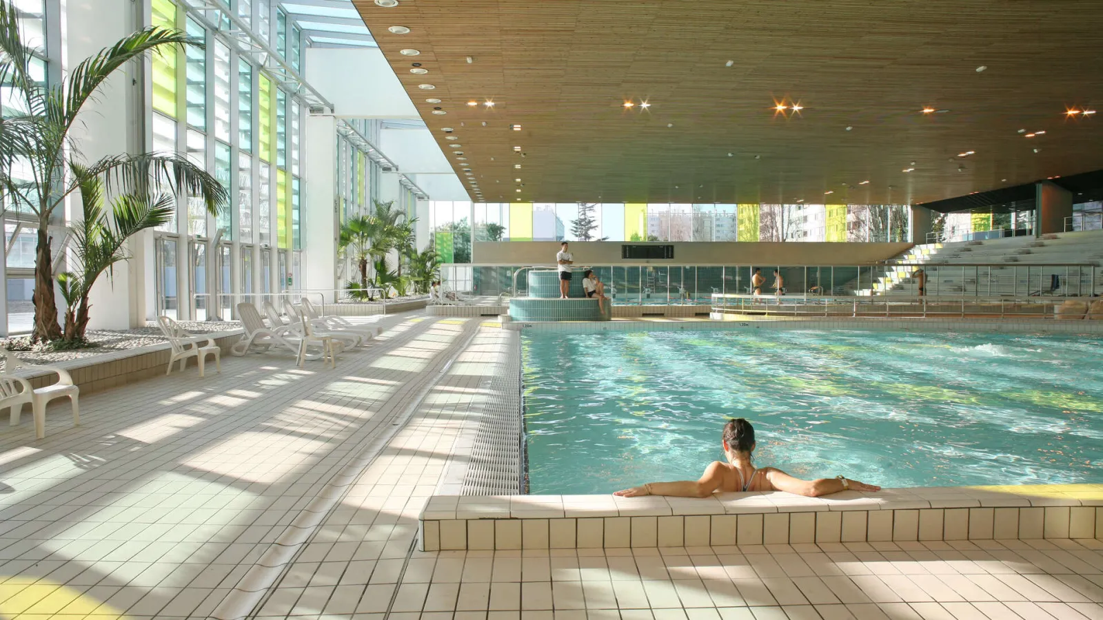 Indoor municipal swimming pool at Grand Parc in Bordeaux featuring an Olympic-sized pool, swimmers, lounge chairs, palm trees, and modern glass architecture.