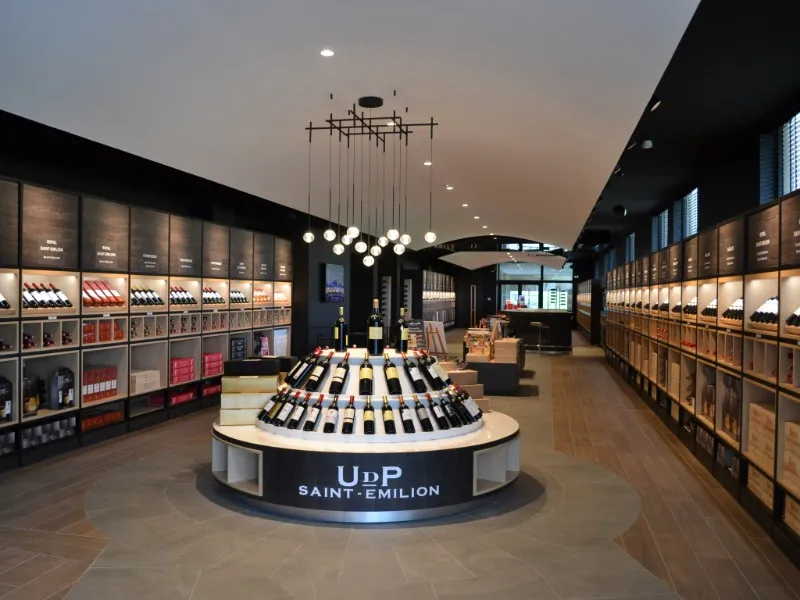 Modern interior of a Saint-Emilion wine tasting cellar in Bordeaux, featuring a circular central display showcase with wine bottles, illuminated glass cabinets on the sides, and a designer ceiling fixture.