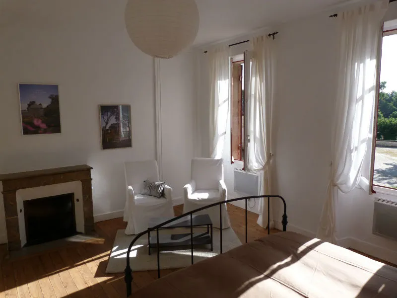 Bright living room at the Maison des Hôtesses in Bordeaux featuring a fireplace, two white armchairs, glass doors opening onto the garden, wooden flooring, and minimalist décor.
