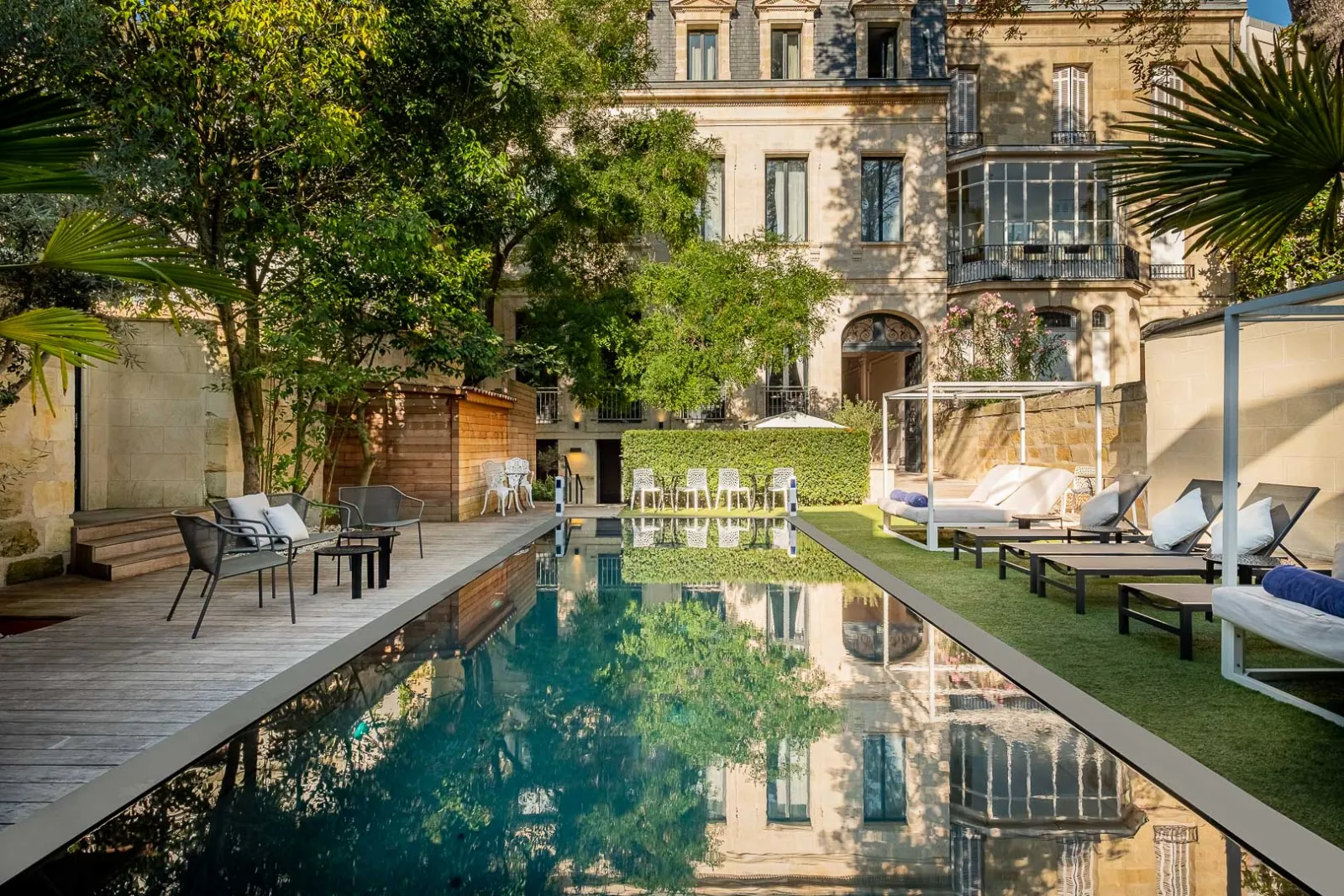 A rectangular reflection pool surrounded by lush green gardens, lounge chairs, and relaxation areas overlooking the elegant classical façade of Palais Gallien Hôtel & Spa in Bordeaux