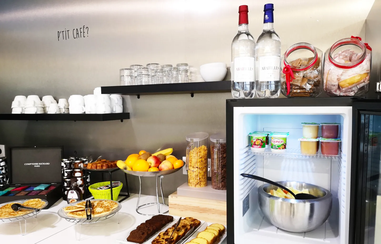 Small café with breakfast counter displaying pastries, fresh fruits, beverages and an open refrigerator with juices and supplies at Mer & Golf City Bordeaux Bassins à Flot apartment hotel.