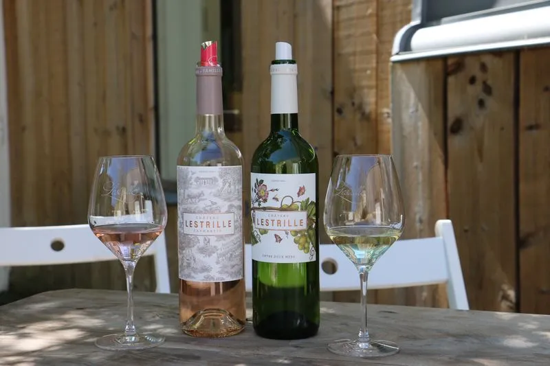 Two wine glasses and two bottles of Château Lestrille, a rosé wine and a white wine, arranged on a wooden table in a rustic setting in Bordeaux.