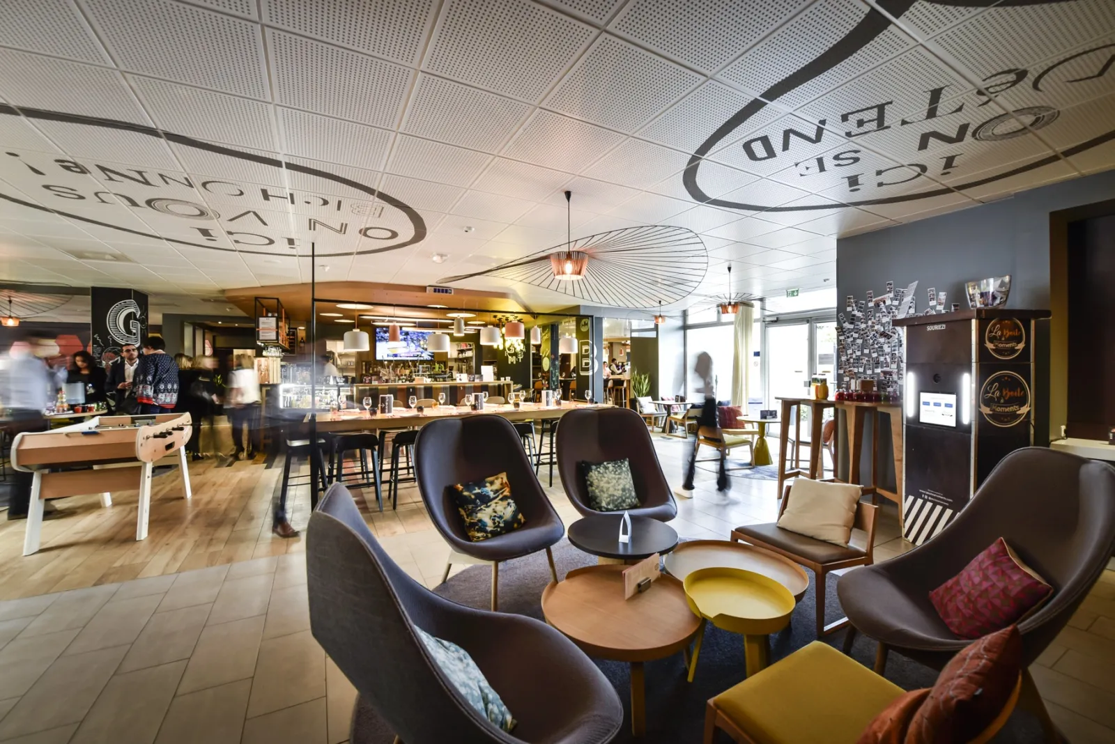 Modern lounge area at Novotel Bordeaux Lac featuring design armchairs, central bar, perforated ceiling, and contemporary décor in grey, black, and ochre tones
