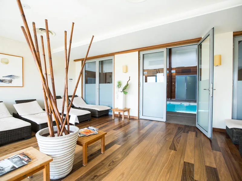 Luxury spa area featuring two black lounge chairs, large glass doors opening to a turquoise indoor pool, wooden floor, and minimalist décor with white vase and bamboo.