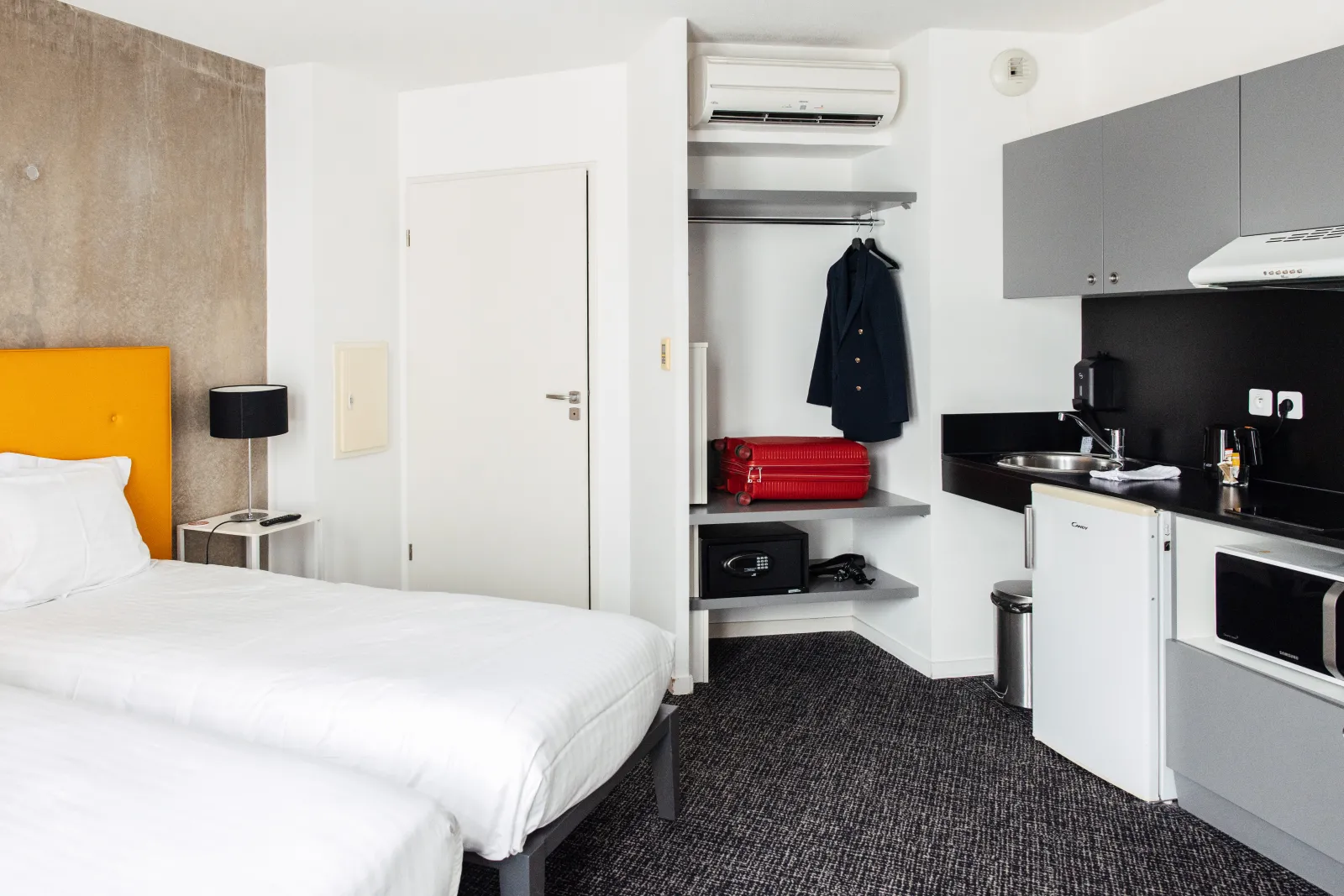 Modern studio apartment at a hotel in Talence, Bordeaux, featuring a white bed with yellow headboard, equipped kitchenette, storage shelves, and air conditioning in minimalist contemporary design