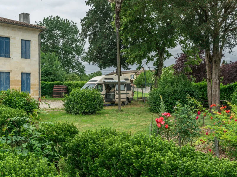 Motorhome parked in the garden of Château Beau Rivage near Bordeaux, surrounded by lush greenery, red flowers, and an old stone building with blue shutters