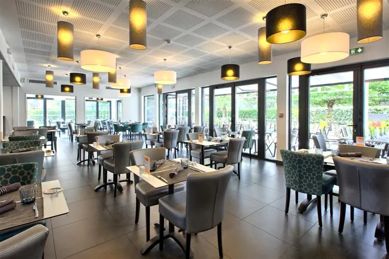 Modern restaurant interior with set tables, gray chairs, designer lighting fixtures, large glass doors opening onto an outdoor terrace with nature views, located in Comté d'Ornon, Bordeaux.