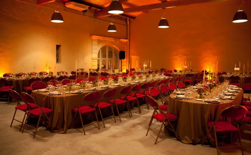 Elegant reception hall at Château Paveil de Luze in Bordeaux, bathed in warm orange-golden lighting, featuring round tables with gold linens, red chairs, place settings, and red-and-pink floral arrangements, beneath exposed ceiling beams and modern pendan