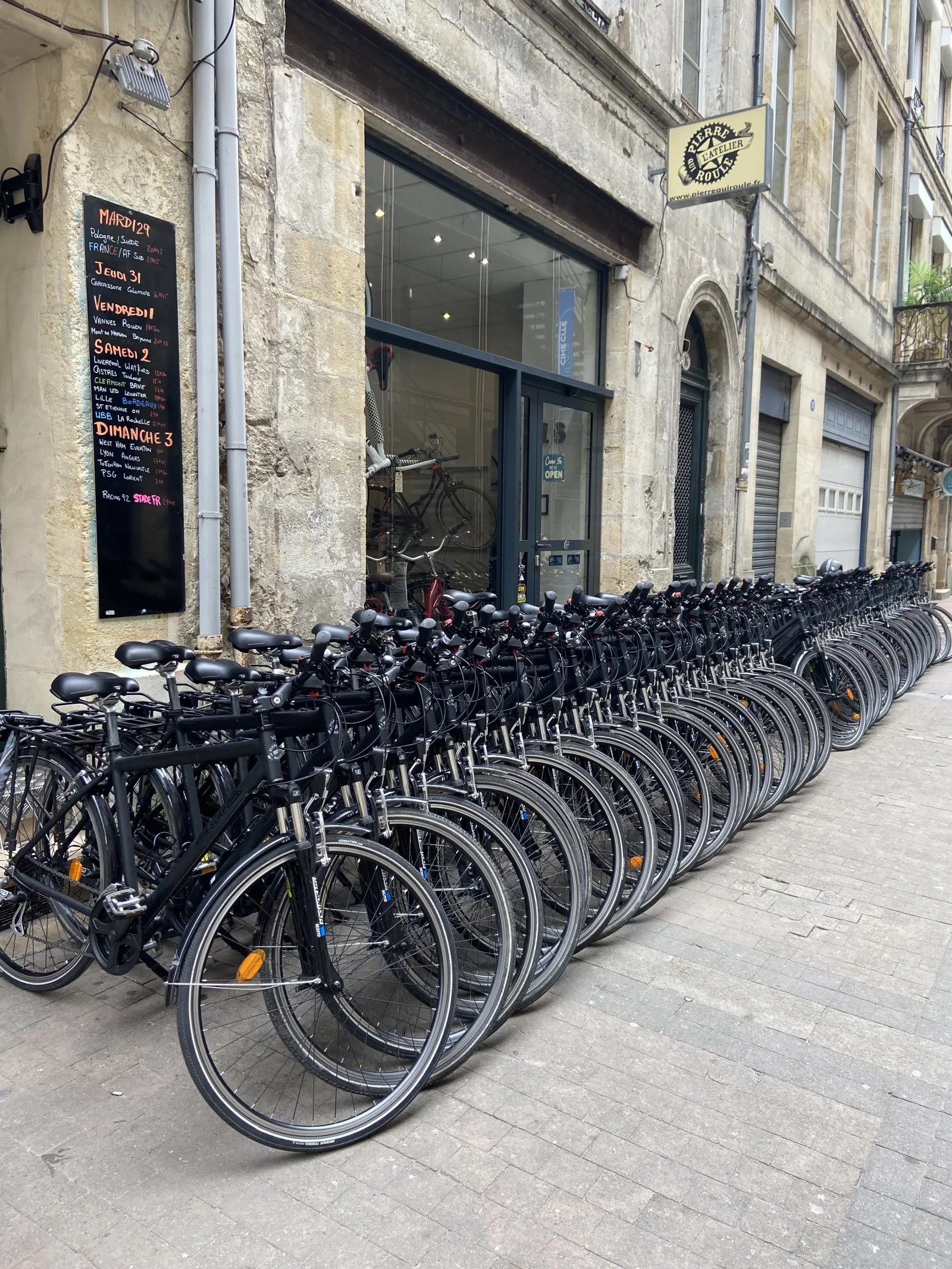A row of black bicycles from Pierre qui Roule bike station lined up along a paved street in Bordeaux, in front of an old building facade with a storefront.