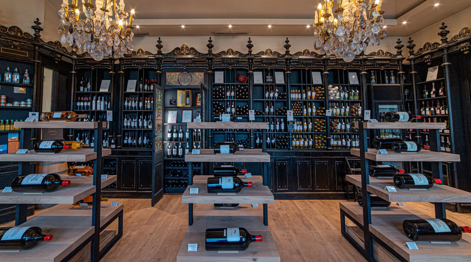 Elegant wine cellar interior with ornate black furniture, wooden tables displaying wine bottles, glass-fronted cabinets filled with bottles, and crystal chandeliers hanging from the ceiling.