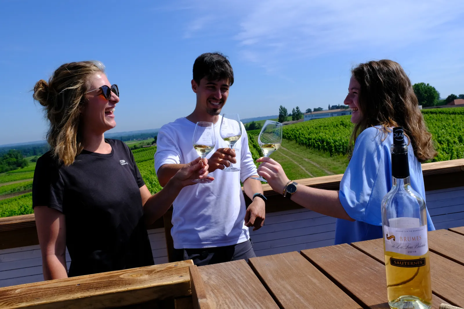 Three people enjoying white wine tasting on a terrace overlooking Bordeaux vineyards at Château La Tour Blanche