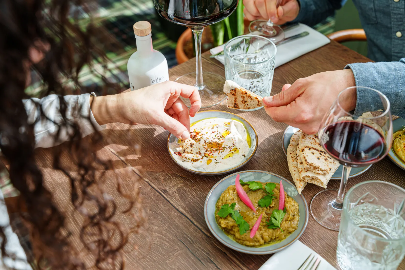 Restaurant diners sharing Mediterranean mezze dishes, toasted bread, red and white wine glasses, and a white spirit bottle on a wooden table.
