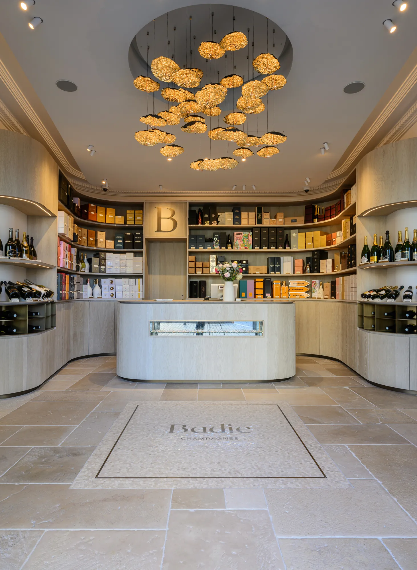 Elegant interior of Badie boutique in Bordeaux featuring a curved white counter, light wood shelving displaying luxury products, and a golden cloud-shaped chandelier on the ceiling.