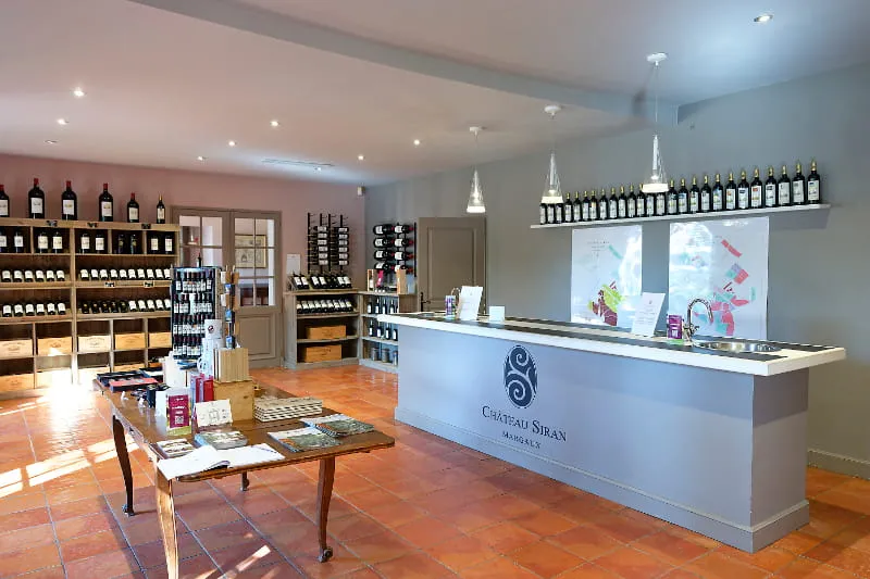 Interior of Château Siran's shop in Bordeaux, featuring a white reception counter, wine bottle shelves, display tables, and modern lighting.