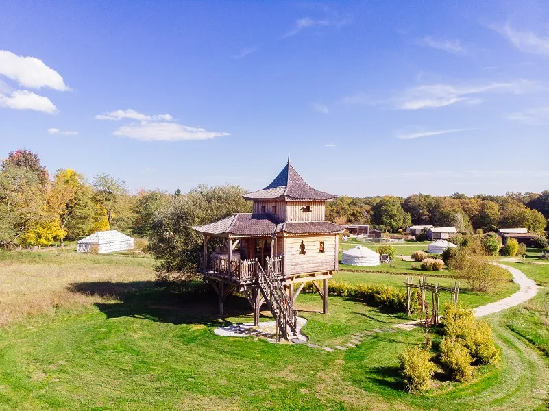 Traditional Asian-style elevated house with pagoda roof in a green park setting at EcÔtelia Estate near Bordeaux, surrounded by autumn foliage and meadows.