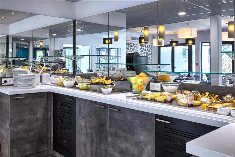 Modern breakfast buffet with black and white counter, fresh fruits, pastries and beverages in a bright room with mirrors and designer lighting at Hôtel Au Comté d'Ornon in Bordeaux.